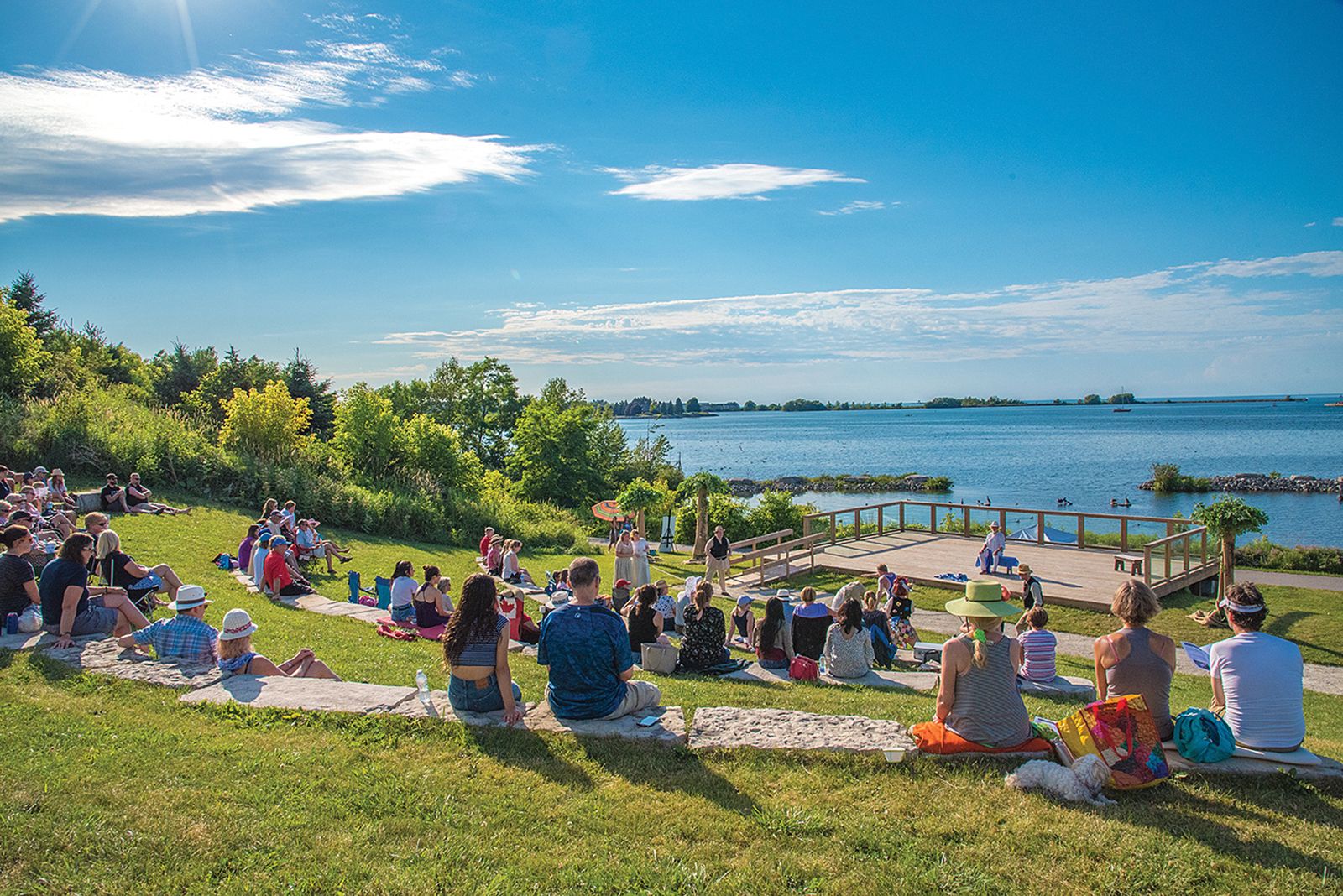 A diverse crowd enjoys a live performance on a sunny day by the Chesapeake's water, surrounded by lush greenery and a clear blue sky.