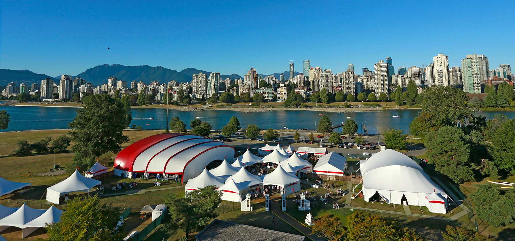 A vibrant outdoor festival setting features large white and red tents, surrounded by lush greenery and a view of a modern city skyline against a clear blue sky.