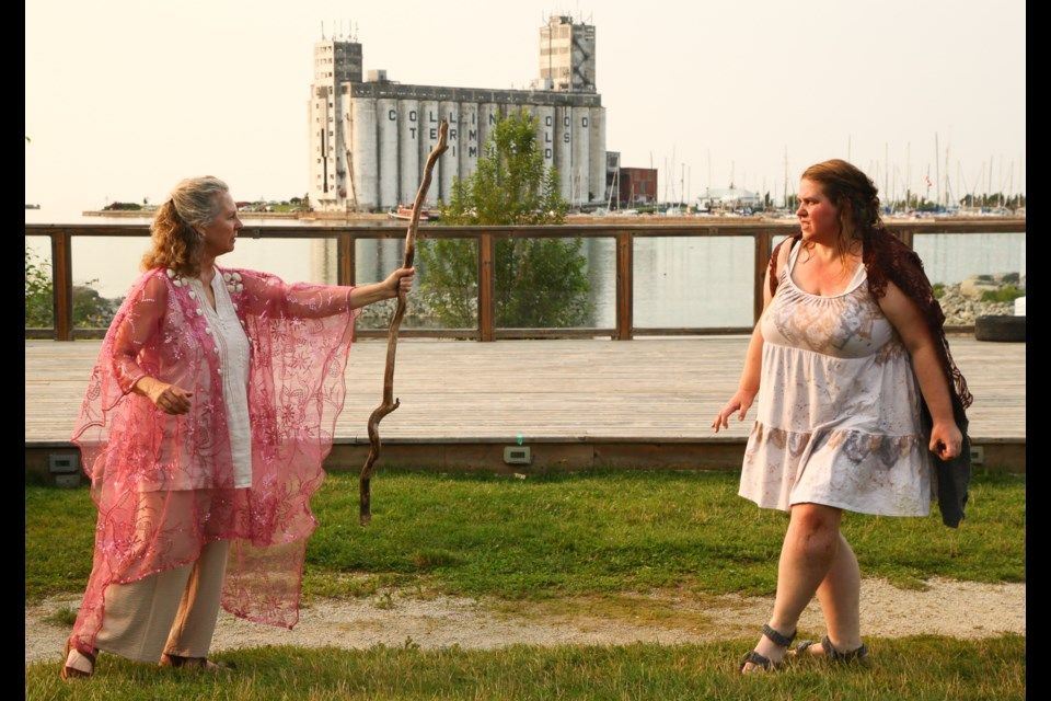 A woman in a flowing pink garment wields a stick as she approaches another woman in a casual dress, with a marina and an industrial building visible in the background.