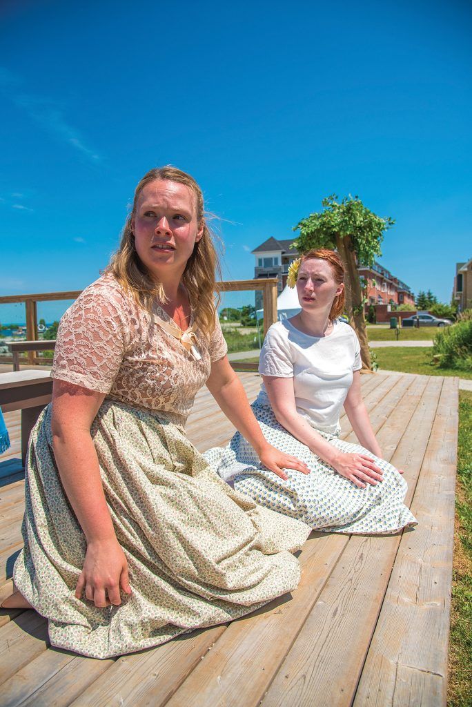 Two women in vintage-style dresses are seated on a wooden deck under a bright blue sky, appearing engaged in a thoughtful conversation as they look out towards the bay.