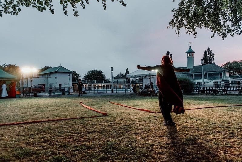 A performer in a flowing cape gestures dramatically on a grassy area marked with red ribbons, illuminated by stage lights and surrounded by a gathering audience at dusk.