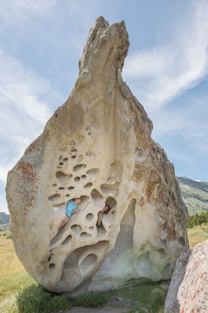 Two people are playfully positioned within a large, uniquely shaped rock that has numerous hollowed-out holes, set against a backdrop of a clear sky and distant mountains in Idaho.