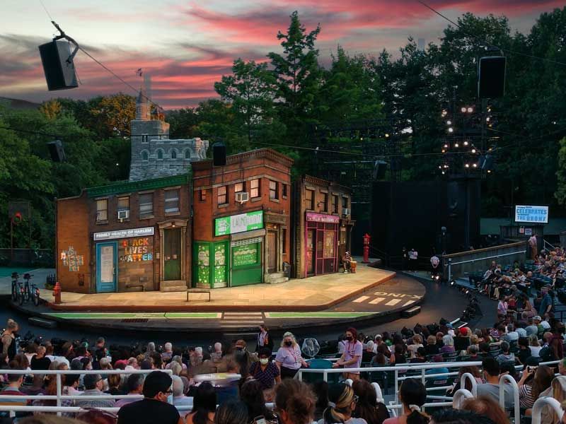 A vibrant outdoor theater set reminiscent of a city street features buildings adorned with colorful signage, as a captivated audience gathers under a stunning sky at dusk.