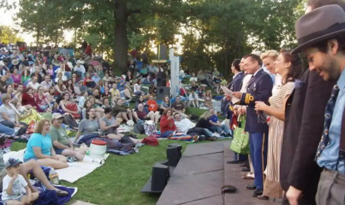 A diverse crowd gathers on blankets in a park, watching performers