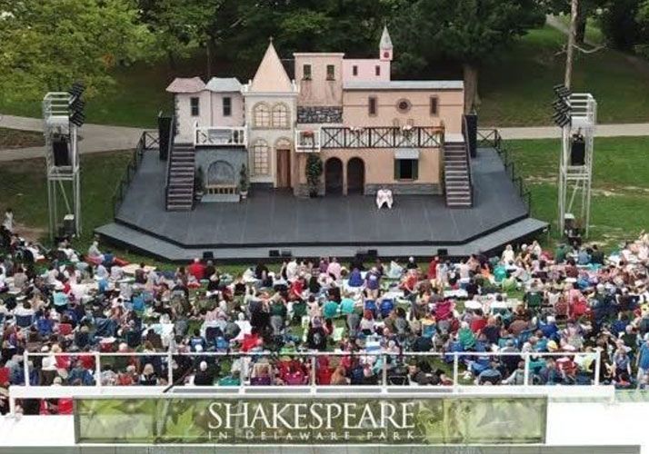 A large audience sits on the grass in front of a Shakespearean stage set