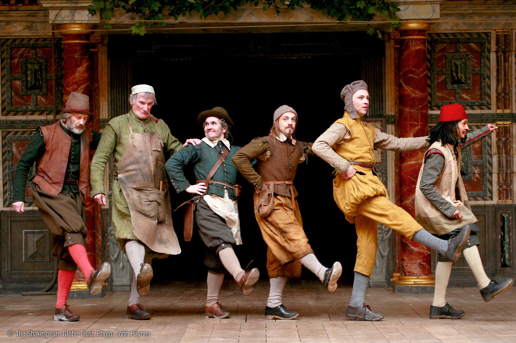 A group of actors in colorful Renaissance costumes are playfully dancing in a line, showcasing their characters' lively energy in a theatrical setting reminiscent of Shakespearean performances.