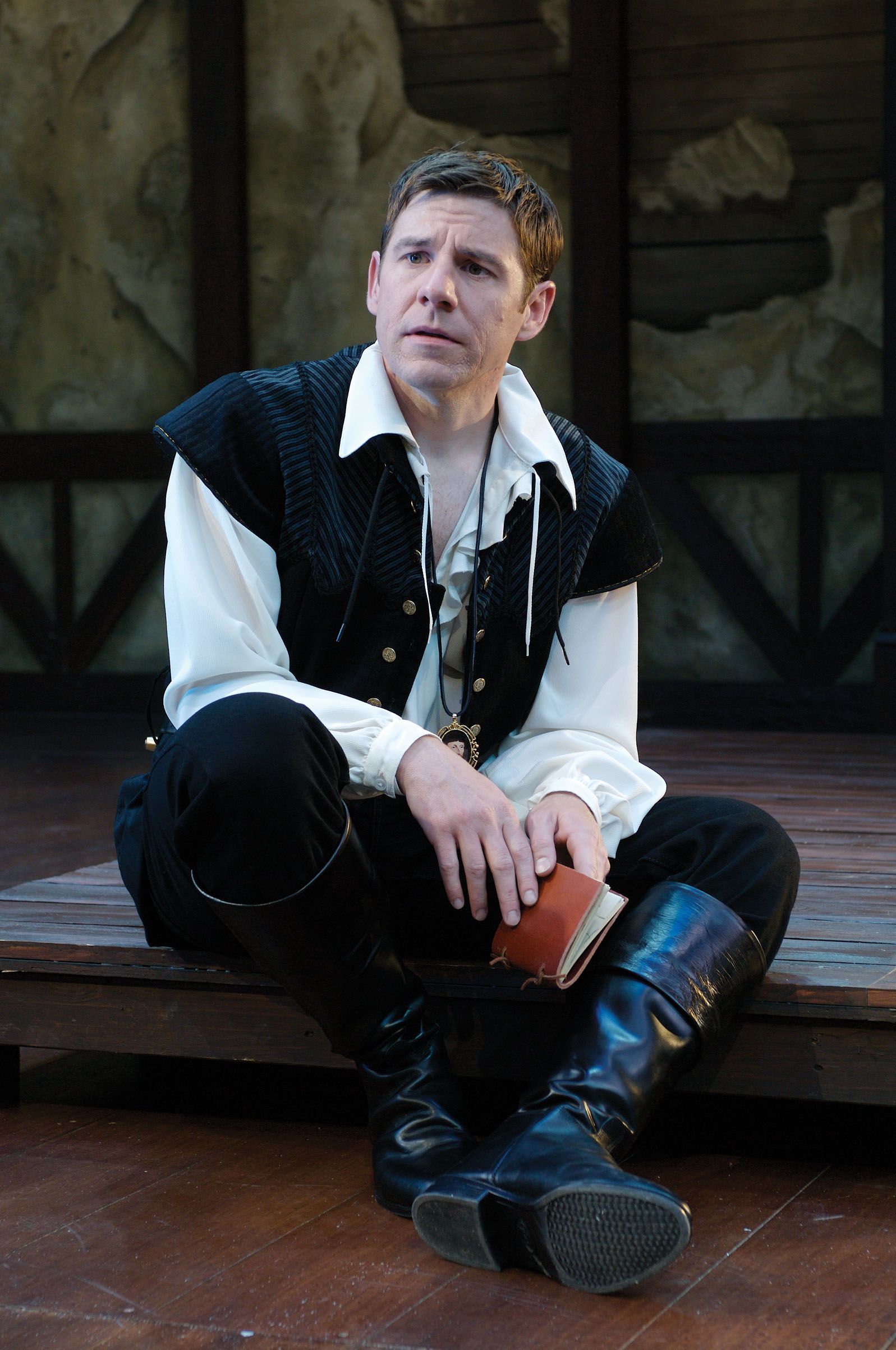 A man dressed in a historical costume, complete with a black vest and tall boots, sits pensively on a wooden platform while holding a small book, conveying a sense of contemplation.