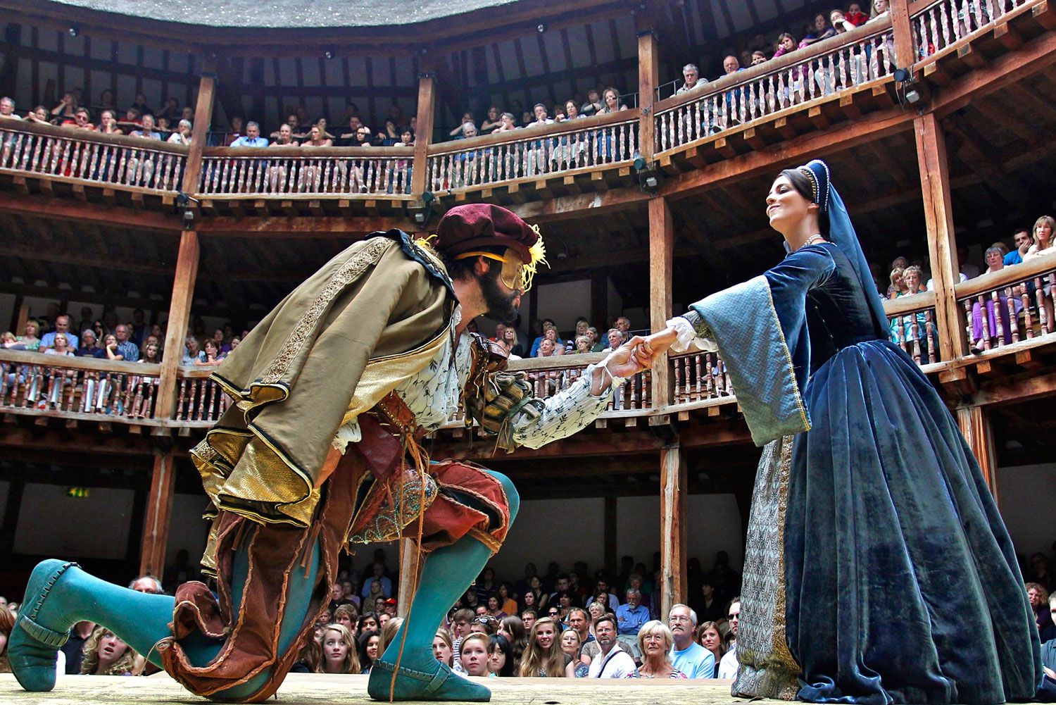 A male actor in elaborate period costume kneels to grasp the hand of a elegantly dressed female actor while an audience watches intently from the surrounding wooden tiered seating.