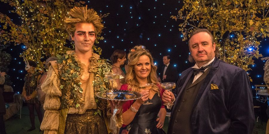 Three elegantly dressed individuals toast with drinks, one in a whimsical leafy costume resembling a character from a Shakespearean play, while the backdrop is adorned with twinkling lights and autumn foliage.