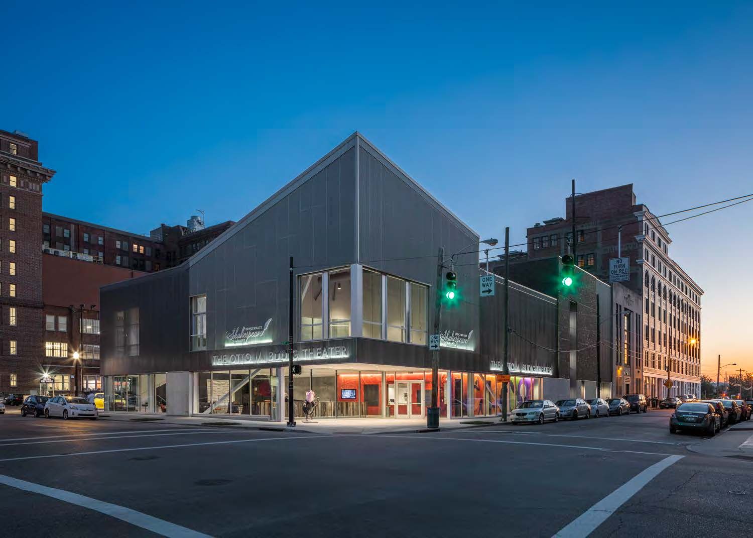 A modern theater building with a distinctive angular design and illuminated signage, located at a busy intersection in Cincinnati, stands in the twilight, surrounded by parked cars and city architecture.