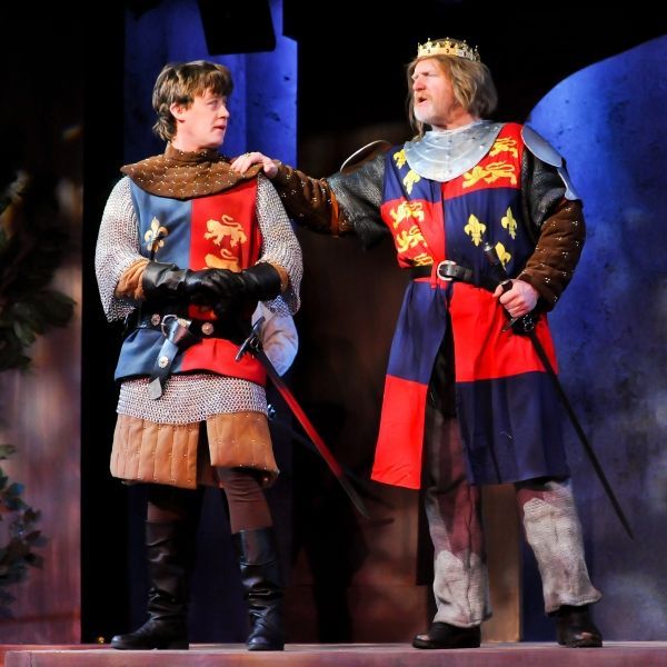 Two actors dressed in medieval costumes, one in a tunic adorned with lions and the other wearing a crown and a colorful coat with a lion and fleur-de-lis design, appear engaged in a dramatic conversation on a theatrical stage.