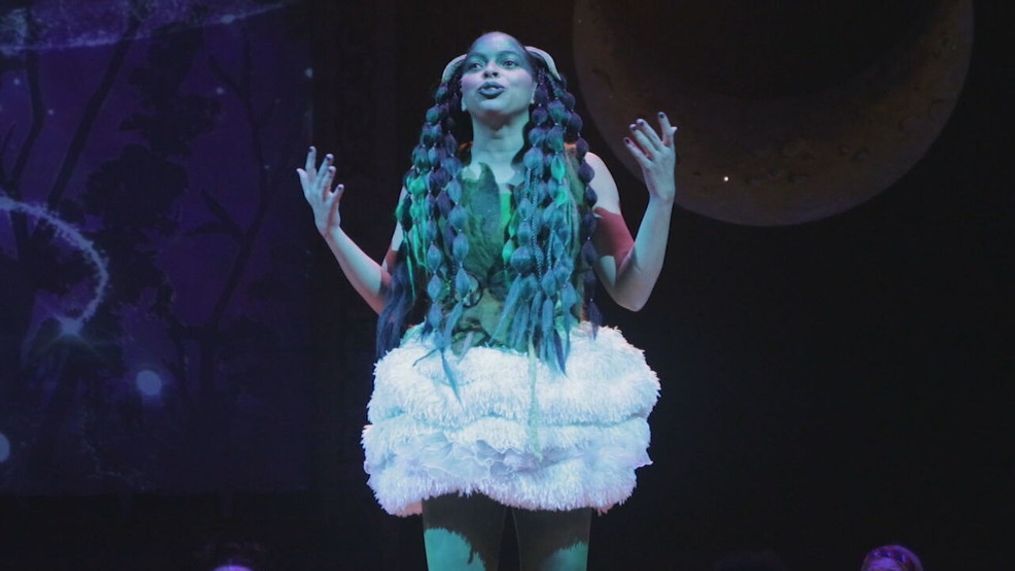 A performer in a whimsical, fluffy costume with vibrant, braided hair passionately engages the audience, set against a backdrop featuring celestial elements.