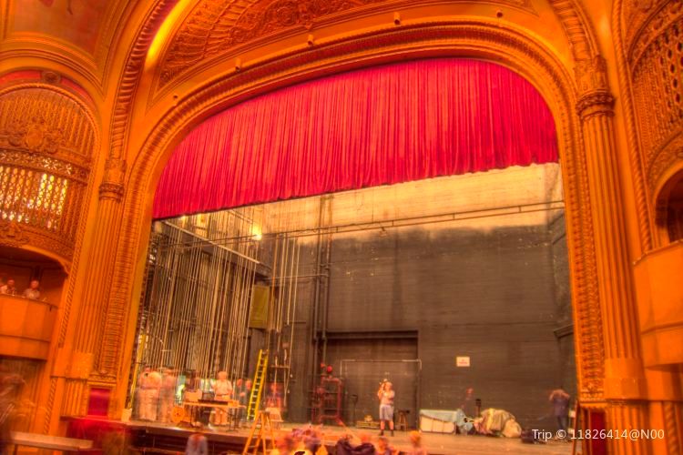 A grand theater stage features a red curtain, ornate golden architecture, and crew members preparing behind the scenes amid various equipment and structures.