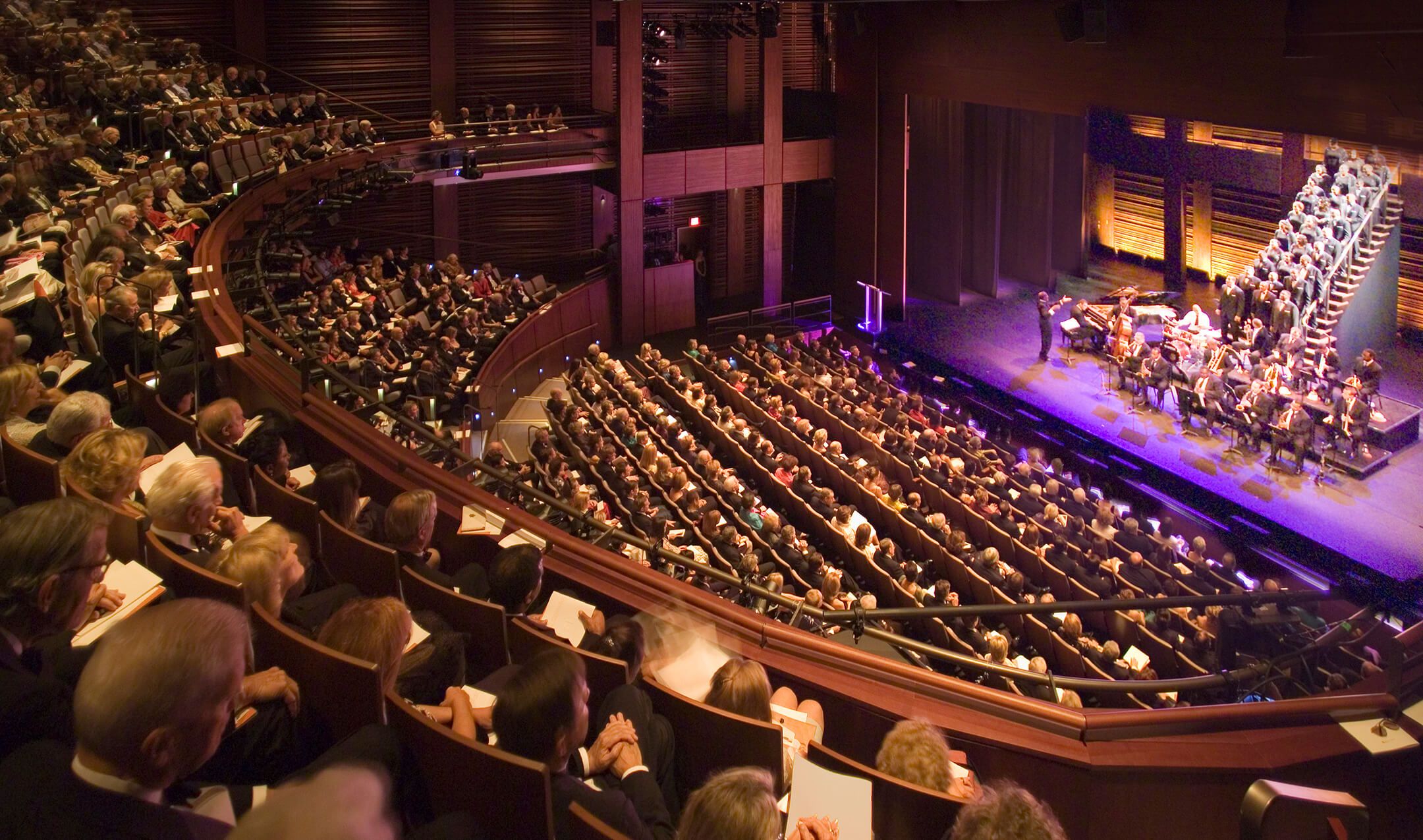 A large audience is attentively watching a performance featuring a musical ensemble on stage, set within a beautifully designed theater space.