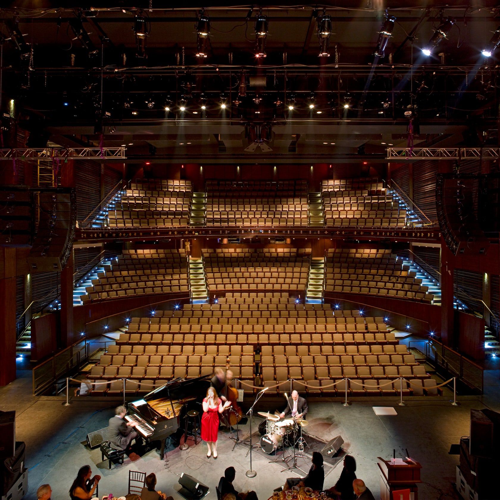 A pianist and a vocalist in a red dress perform on stage in front of an expansive, empty theater with tiered seating and sophisticated lighting.