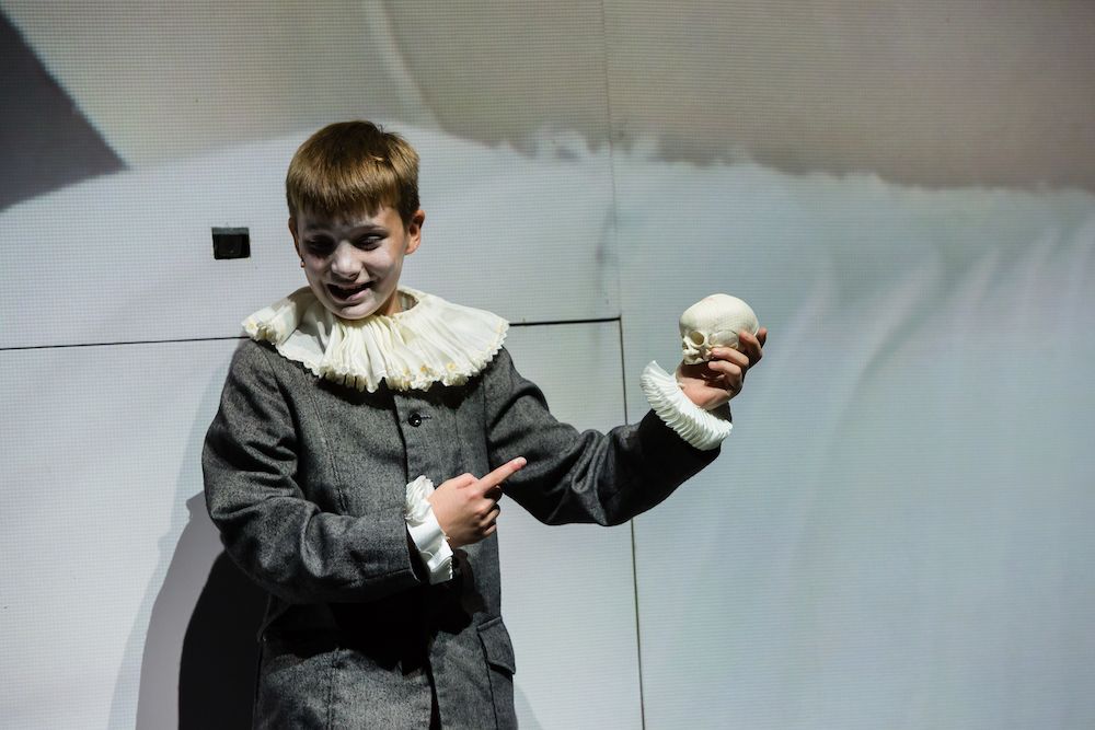 A young boy dressed in period clothing with a ruffled collar holds a small skull and gestures expressively, conveying themes related to Hamnet and Shakespeare's exploration of mortality.