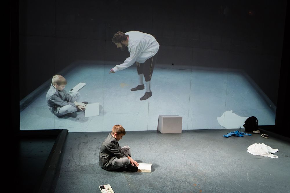 A young boy sits on the ground, focused on a book, while a larger projection of another boy and a man engaged in a conversation looms on a wall behind them, creating a poignant contrast between the two scenes.