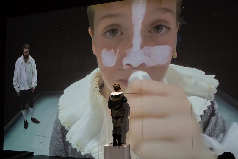 A young boy stands on a pedestal, preparing to apply white face paint, while larger images of a man and another child dressed in period costumes are displayed on the backdrop, suggesting a theatrical context related to "Hamnet."