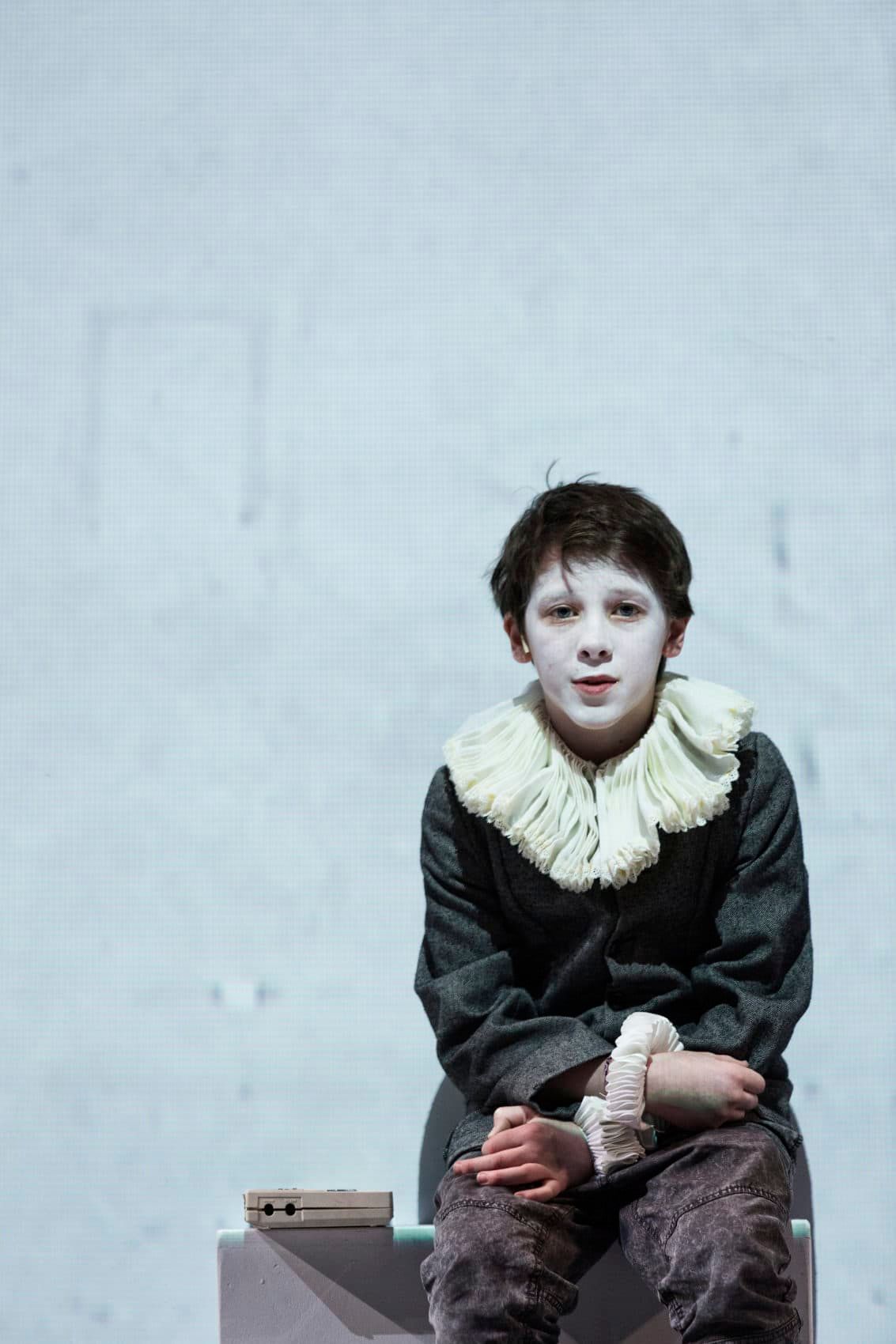 A young boy dressed in period clothing with white face paint and a ruffled collar sits on a stool, expressing a contemplative demeanor against a muted backdrop.