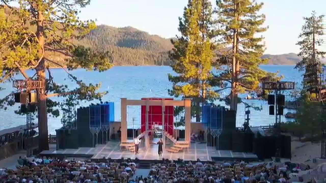 A stage is set by a picturesque lake in Tahoe, surrounded by tall trees, as an audience gathers to watch a Shakespearean performance.