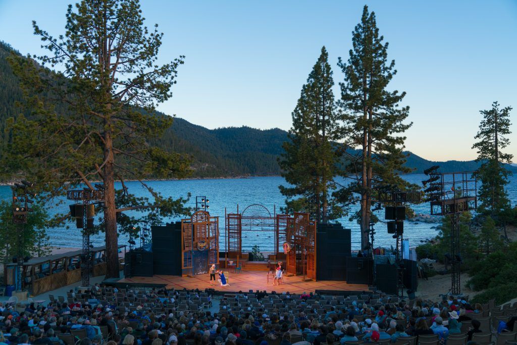 A theatrical performance of Shakespeare takes place on a lakeside stage surrounded by tall trees, with an audience seated in rows facing the water.