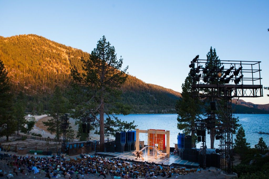 A large outdoor theater filled with an audience is set against a picturesque lakeside backdrop and surrounding mountains, featuring a colorful stage where a performance is taking place.