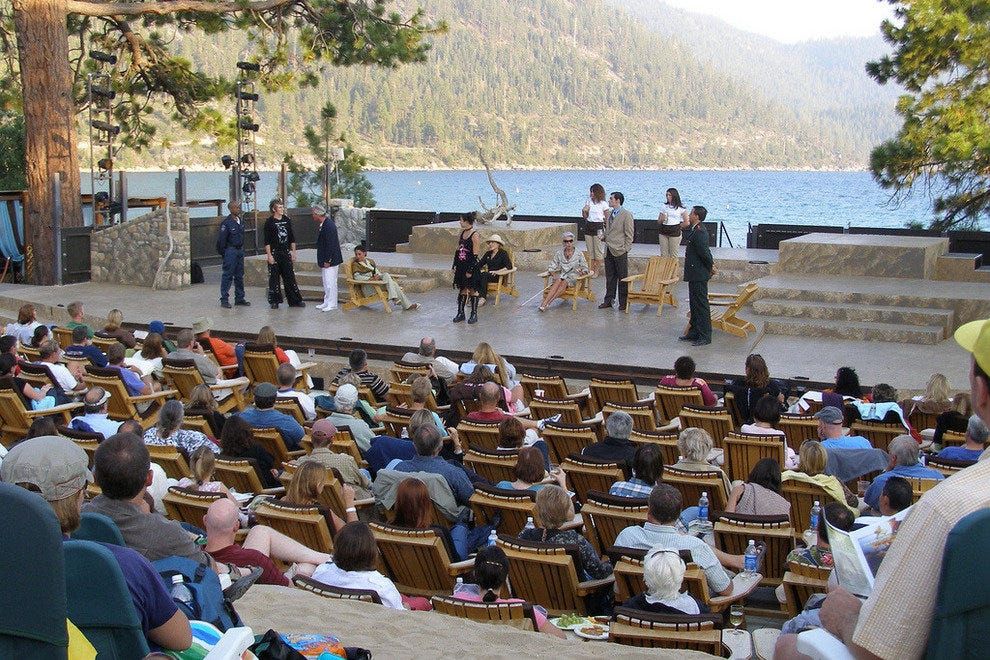 A live performance of Shakespeare is taking place by a lakeside, with actors on stage and an engaged audience seated in wooden chairs, surrounded by trees and mountains in the background.