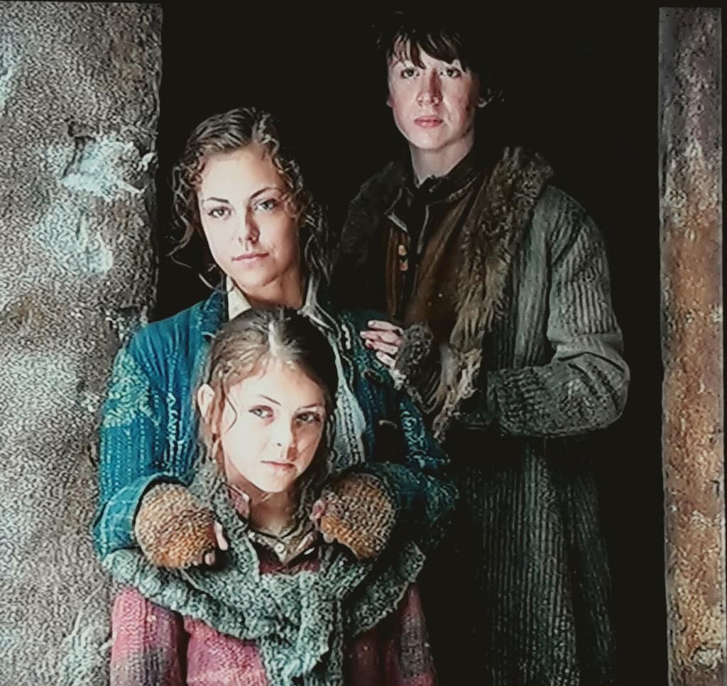 Three siblings stand together in a dimly lit space, with one older brother behind, while the two sisters in front wear expressions of curiosity and resilience, dressed in warm, layered clothing.