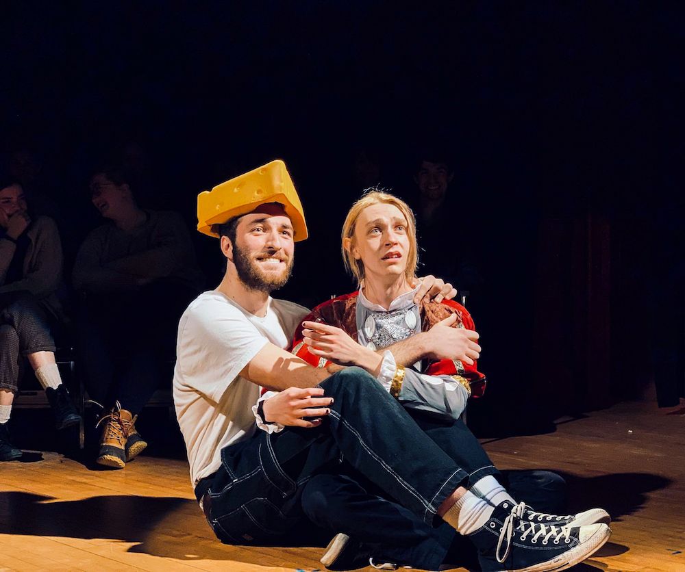 Two actors are seated close together, one wearing a comical cheese hat