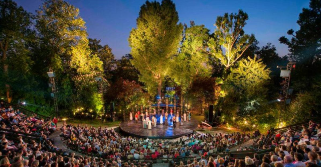 A vibrant outdoor theater filled with an engaged audience watching a performance that blends teenage emotions and Shakespearean humor, surrounded by lush greenery and illuminated by soft stage lights.