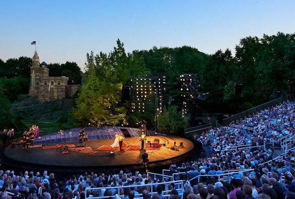 A vibrant outdoor theater production is taking place at dusk with a large audience gathered, lush greenery surrounding the stage, and a historical building visible in the background.