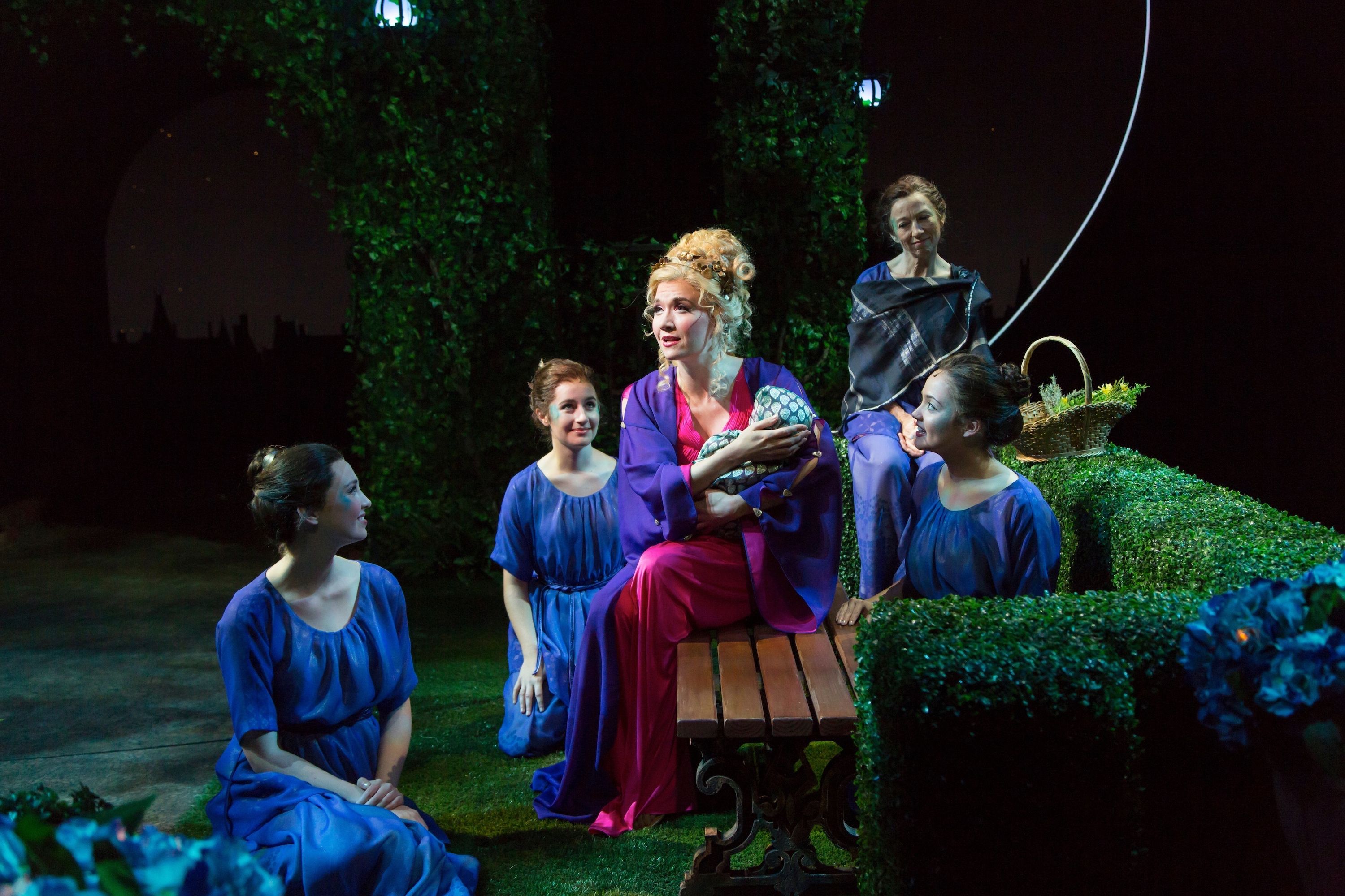 A group of women dressed in flowing gowns sits in a lush, green garden setting, engaging in an animated conversation, with one woman in a vibrant pink dress holding a creature in her arms while the others listen intently.